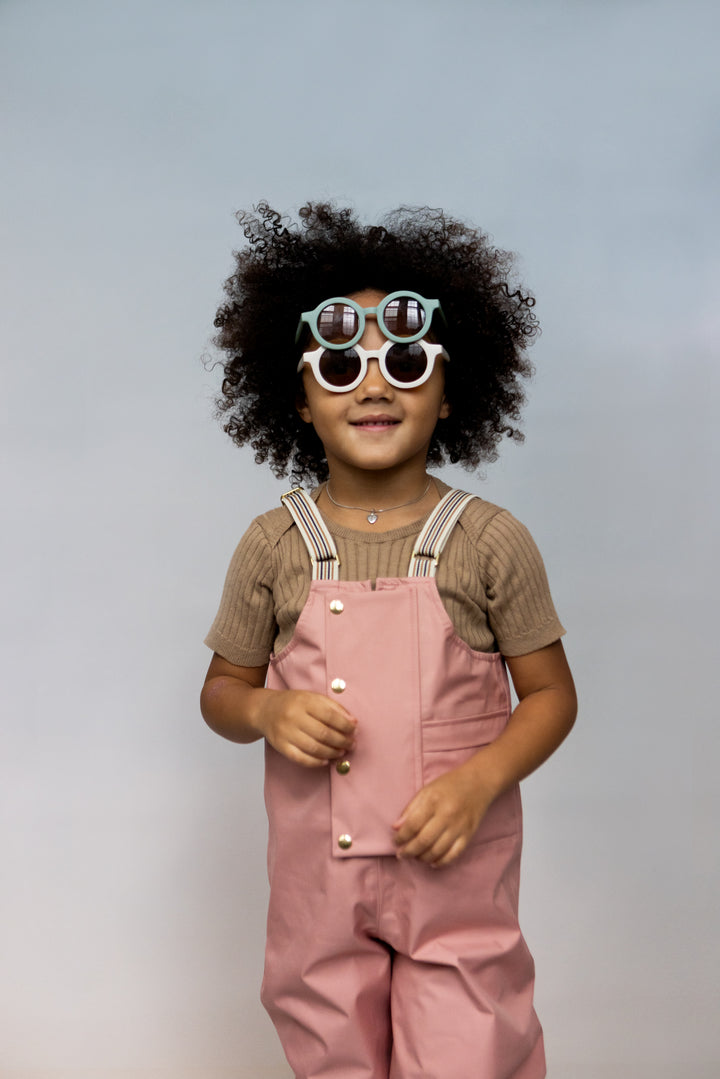 smiling kid with black curly hair and posing with faire child childrens rain dungarees in beach rose enfant posant avec une salopette de pluie imperméable 